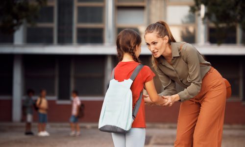 Mid adult mother holding hands with her daughter while greeting with her on first day of school in the schoolyard. Copy space.