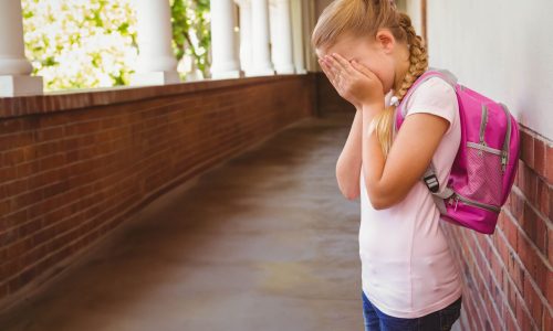 Female child covering face leaning against brick wall in school hall with pink backpack, copy space. Child, corridor, architecture, greenery, classroom, youthful, introspection