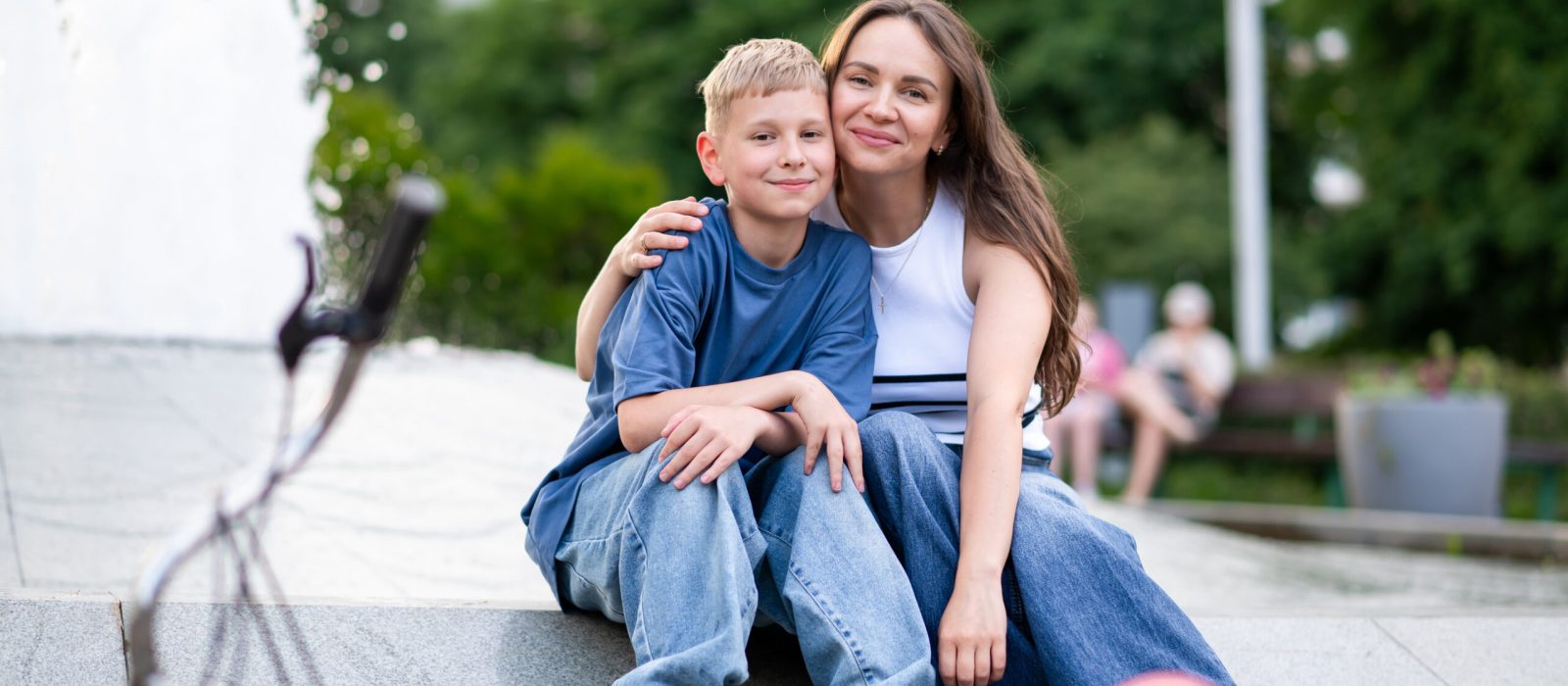 Happy mother and teenage son sitting hugging near fountain in ur