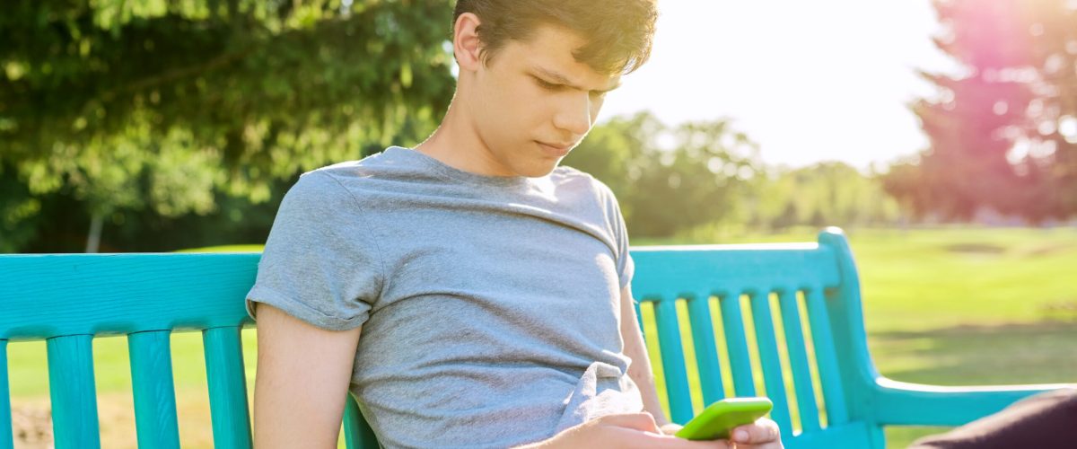 Nastolatek korzysta ze smartfona, siedzi na ławce w parku.