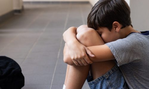 Sitting on floor, boy hugging knees and feeling sad in school hallway. Sadness, bullying, loneliness, depression, anxiety, mental health