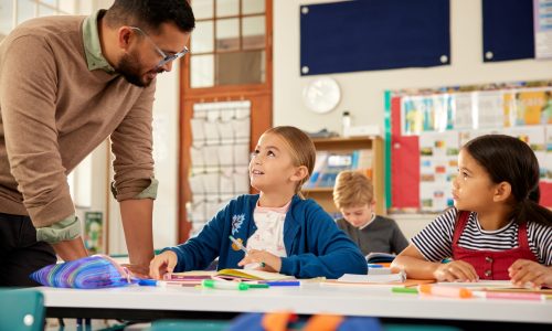 Male teacher in classroom helping multiethnic kids for studying at desk. Little schoolgirls in classroom asking for help with homework. Confident teacher helping elementary students in understanding lesson at school.